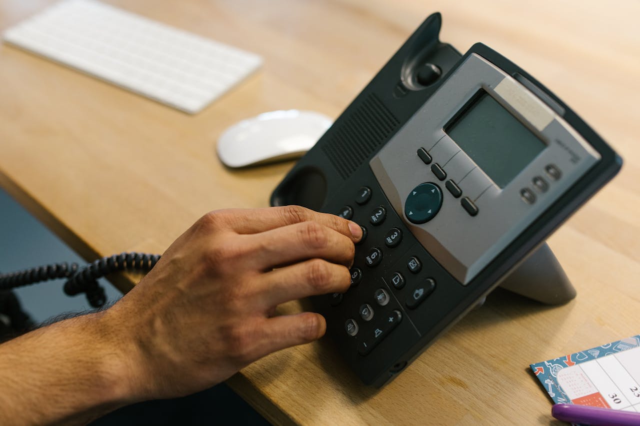 who-we-are A hand dialing a phone on a wooden desk. Office environment with keyboard and planner.